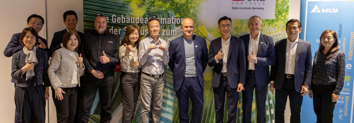 Gruppe von Geschäftsleuten und Mitarbeitern bei Delta Controls Germany, die die Übernahme feiern, vor einem grünen Hintergrund mit Delta-Logo.