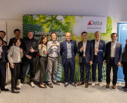 Gruppe von Geschäftsleuten und Mitarbeitern bei Delta Controls Germany, die die Übernahme feiern, vor einem grünen Hintergrund mit Delta-Logo.